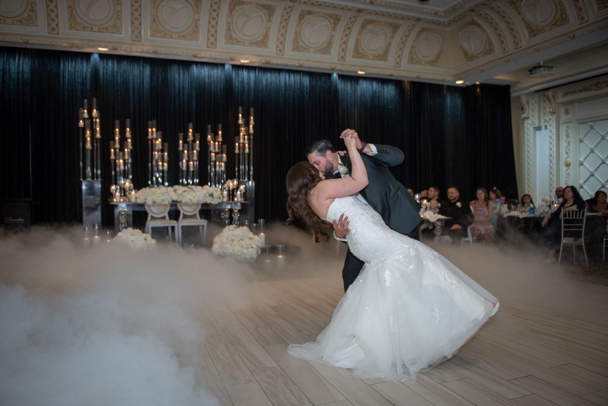 Bride and groom dance intimately in swirling fog on wooden floor, surrounded by tall candelabras and seated guests.