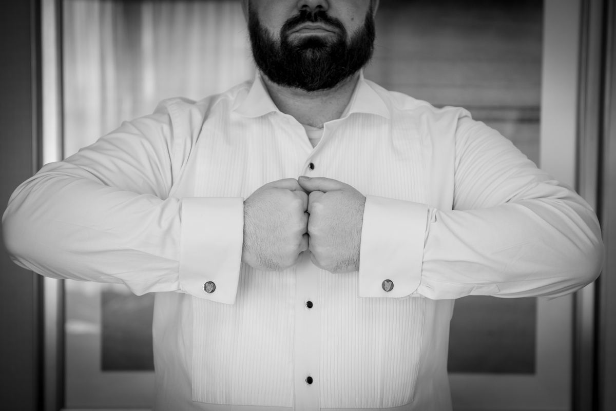 A man with a beard adjusts his pinstriped shirt cuffs, hands clasped at chest, dressed for a formal event.
