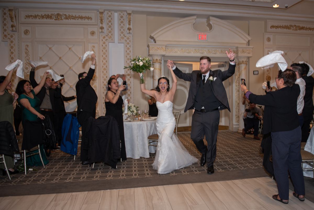 Bride and groom exit hall amid cheering guests showering them with napkins, surrounded by festive celebration.