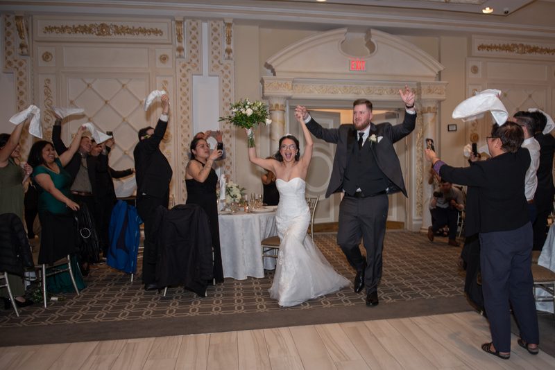 Bride and groom exit hall amid cheering guests showering them with napkins, surrounded by festive celebration.