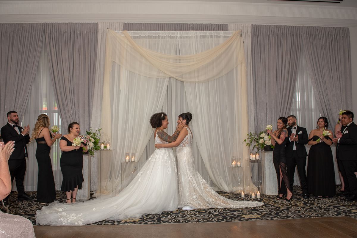 Two brides embrace tenderly at the center of a wedding ceremony, surrounded by applauding guests in formal attire.