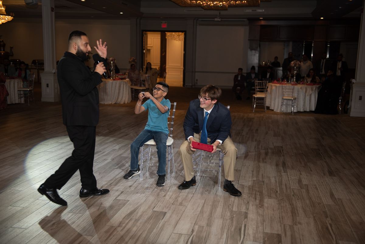 A man in a black suit interacts with two boys on a wooden dance floor, one laughing and the other holding a red box.