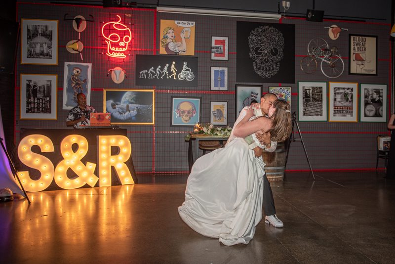 A bride and groom dance intimately at a reception with a vibrant wall display behind them, illuminated by warm lighting.