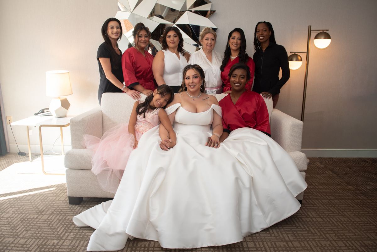 A bride in a white gown sits with bridesmaids and a young girl in a pink tutu in a warmly lit, stylish room.