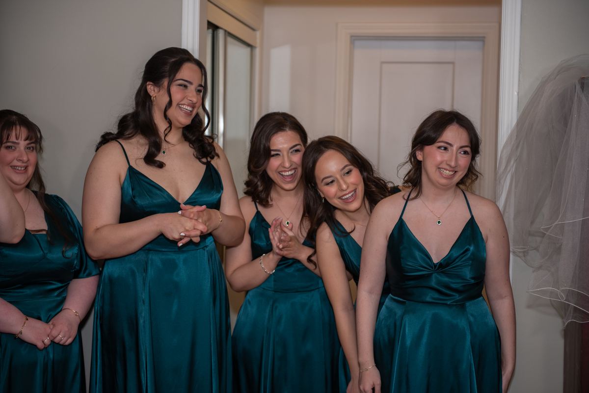 A bride and her bridesmaids in teal satin gowns laugh together, hands clasped, with a veil hanging nearby.