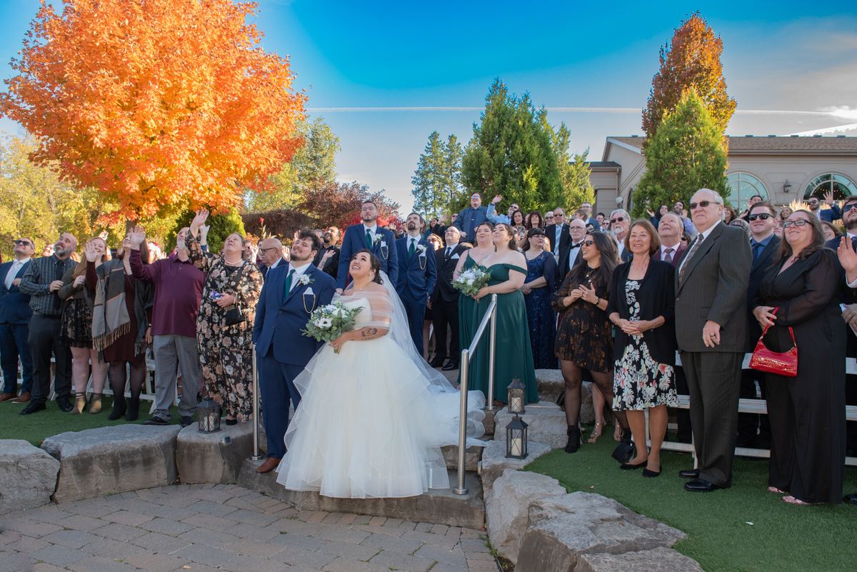 A bride in a white gown stands on stone steps with guests clapping and foliage ablaze in autumn colors.