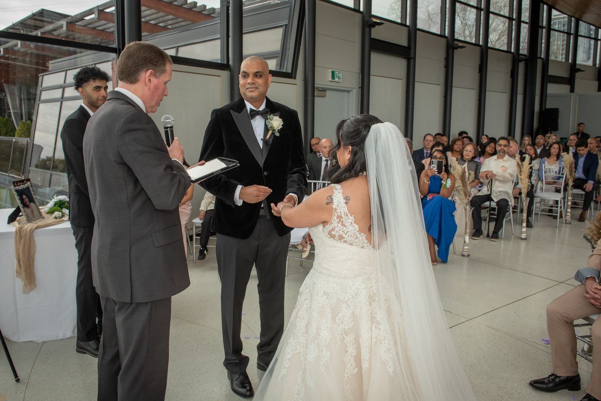 Bride in lace gown and veil holds groom's hand as officiant reads from tablet during indoor ceremony.