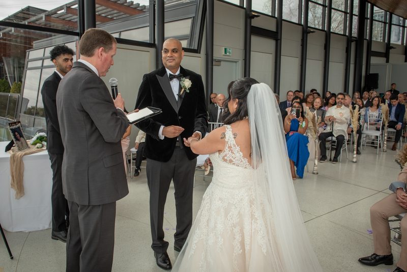 Bride in lace gown and veil holds groom's hand as officiant reads from tablet during indoor ceremony.
