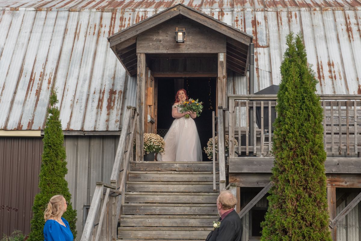 A bride in a white gown smiles from the top of wooden stairs leading to a rustic barn with a metal roof.