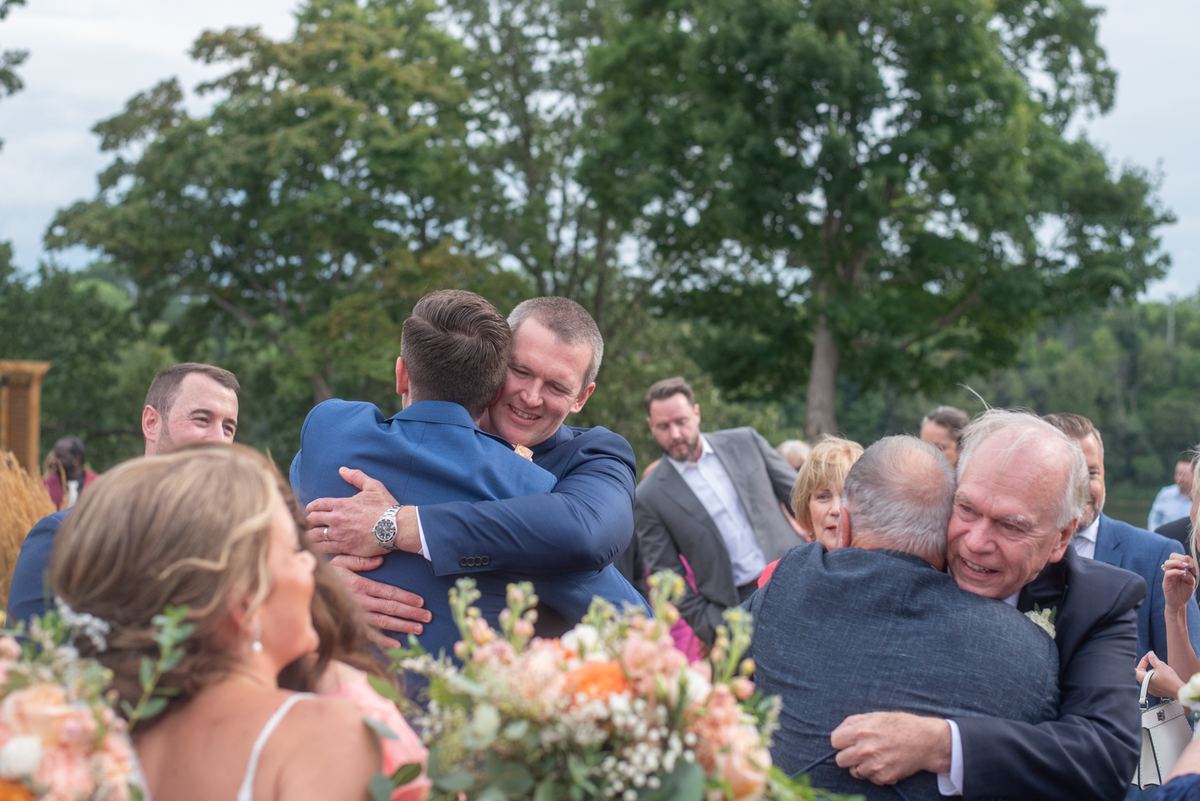 Guests embrace joyfully under trees at an outdoor wedding, surrounded by flowers and dressed in semi-formal attire.
