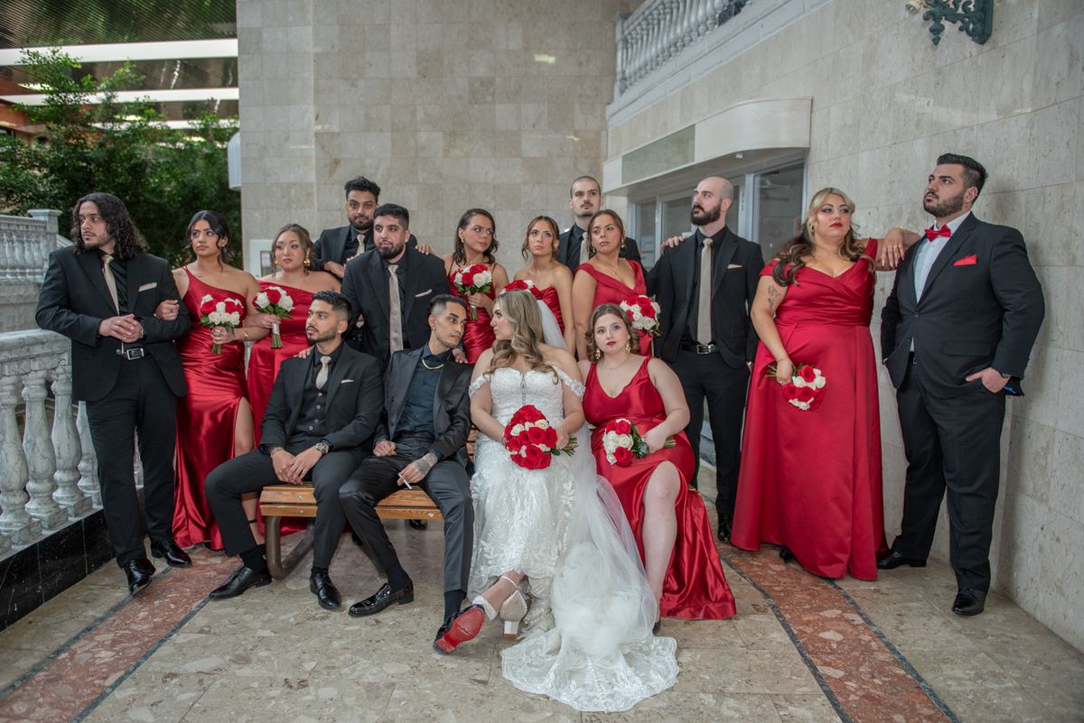 Bride in white gown and groom in black suit with attendants in red satin gowns and black suits holding bouquets in courtyard.