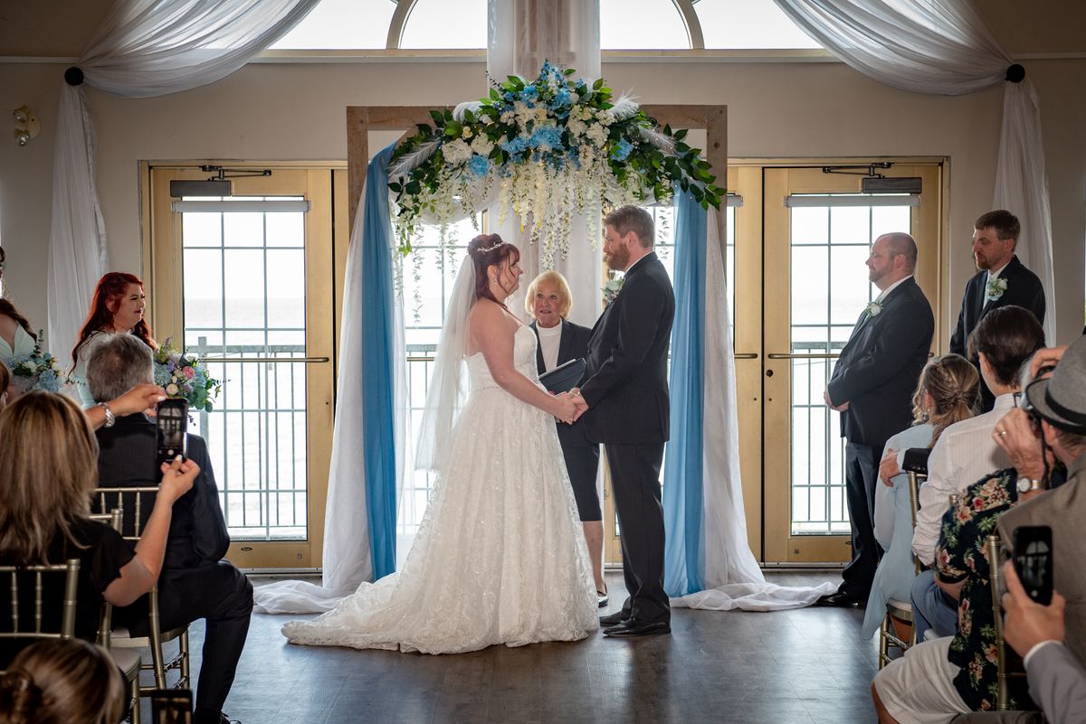 A bride and groom stand under a floral arbor, holding hands as they exchange vows in front of guests seated around them.