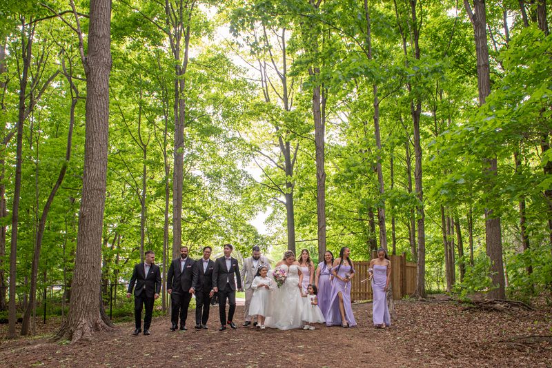 A wedding party poses in a sun-dappled forest clearing, with the bride and groom at center surrounded by attendants.
