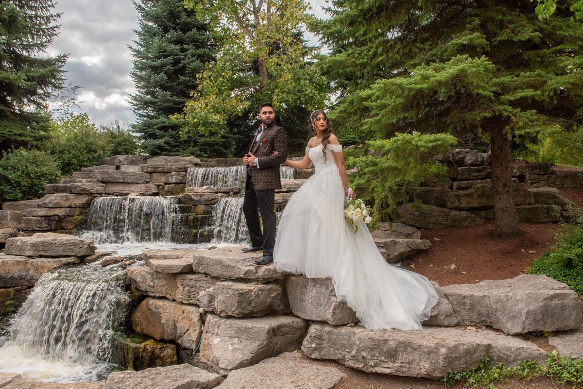 A bride in a white gown and groom in tweed stand on mossy stones beside a cascading waterfall in a forest garden.
