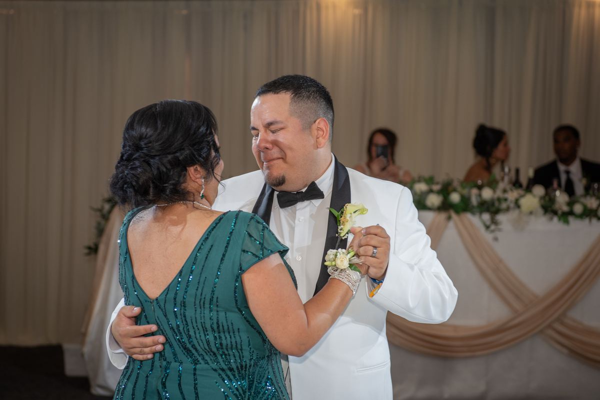 A man and woman in formal attire dance closely, smiling through tears under soft lighting with drapes.