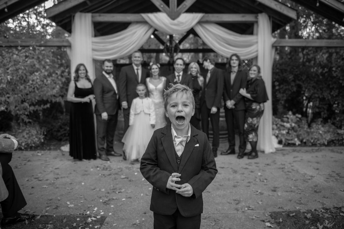 A young boy in a suit shouts with joy during a wedding, interrupting a posed family photo under a wooden arch.