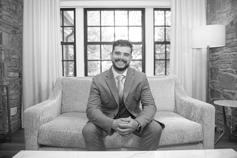 A man in a suit smiles on a textured sofa in a rustic room with large windows, bathed in soft light.