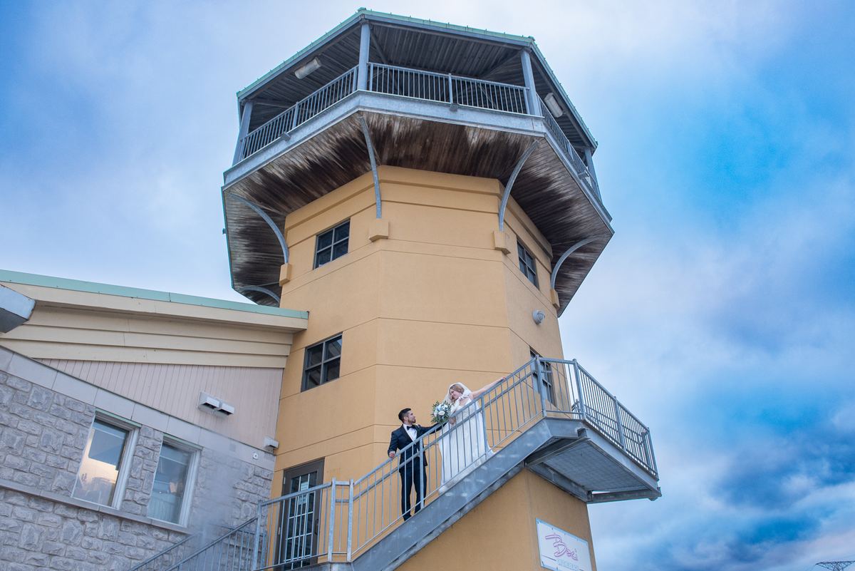 A bride and groom stand on the metal staircase of a yellow tower, smiling against a partly cloudy sky.