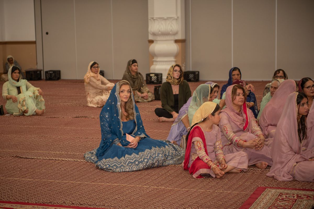 Women and girls in traditional attire sit cross-legged on a carpeted floor in a prayer hall, engaged in quiet reflection.
