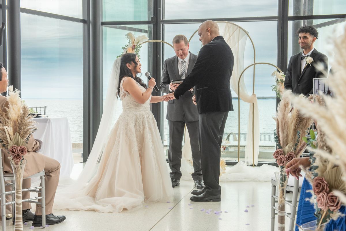 A bride and groom exchange vows by the ocean, surrounded by pampas grass and dried roses under a minimalist gold arch.