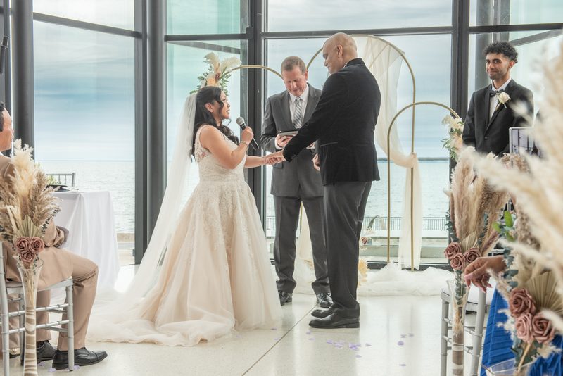 A bride and groom exchange vows by the ocean, surrounded by pampas grass and dried roses under a minimalist gold arch.