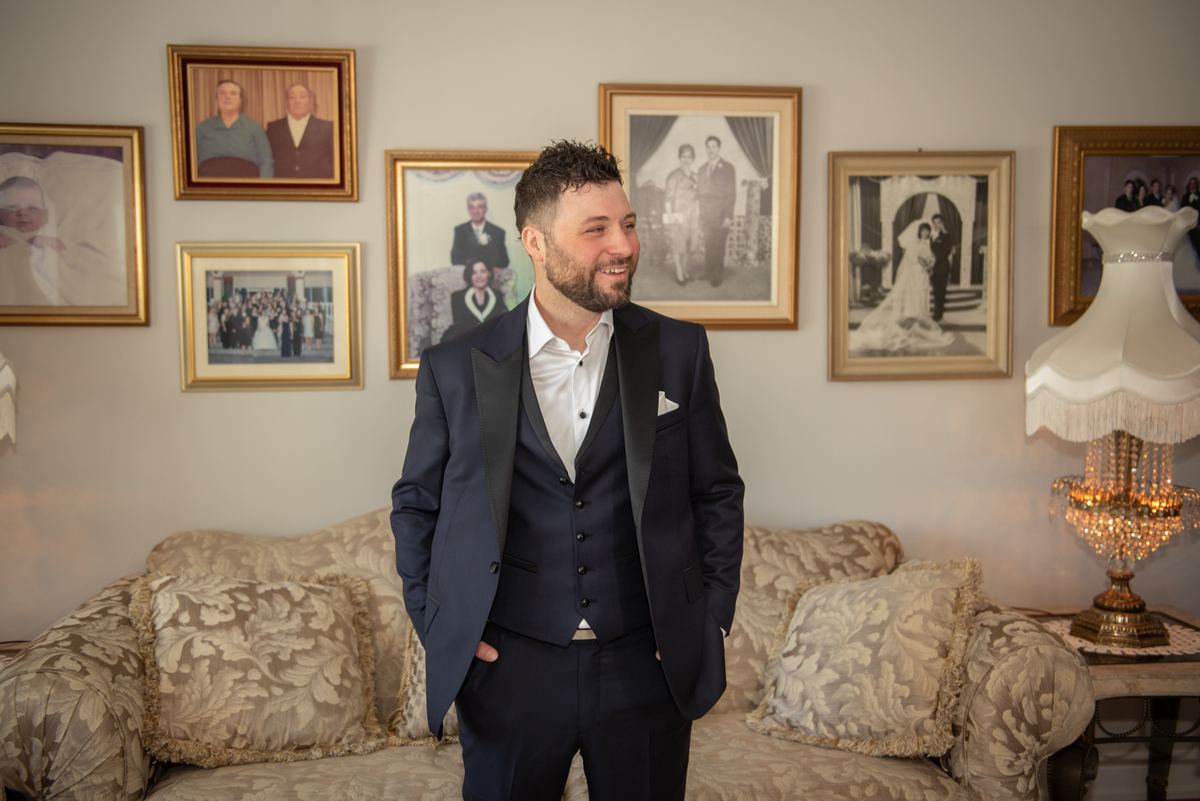 A groom in a navy tuxedo stands smiling in a room with family photos and wedding portraits, illuminated by an ornate lamp.