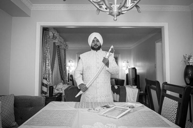 A Sikh groom in white attire holds a raised kirpan in a dimly lit dining room decorated for a ceremony.