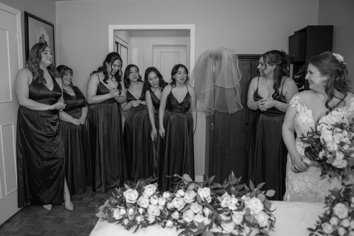 A bride in a lace gown and veil stands with her smiling bridesmaids in matching satin gowns, sharing a joyful moment.