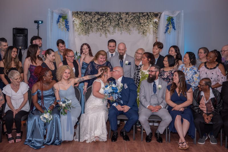 Jennifer and Joe share a tender kiss at their wedding, surrounded by seated guests in festive attire.
