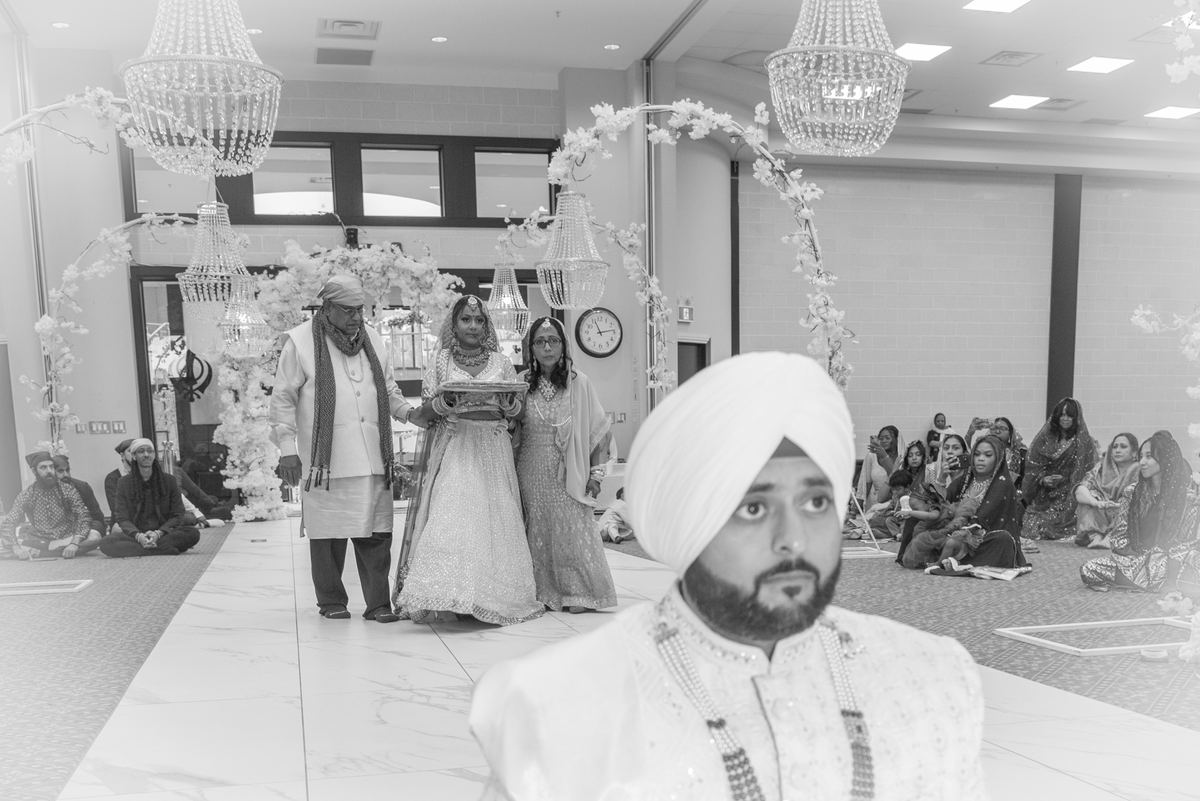 A Sikh groom in white turban stands at the forefront during wedding ceremony under floral arches and chandeliers.