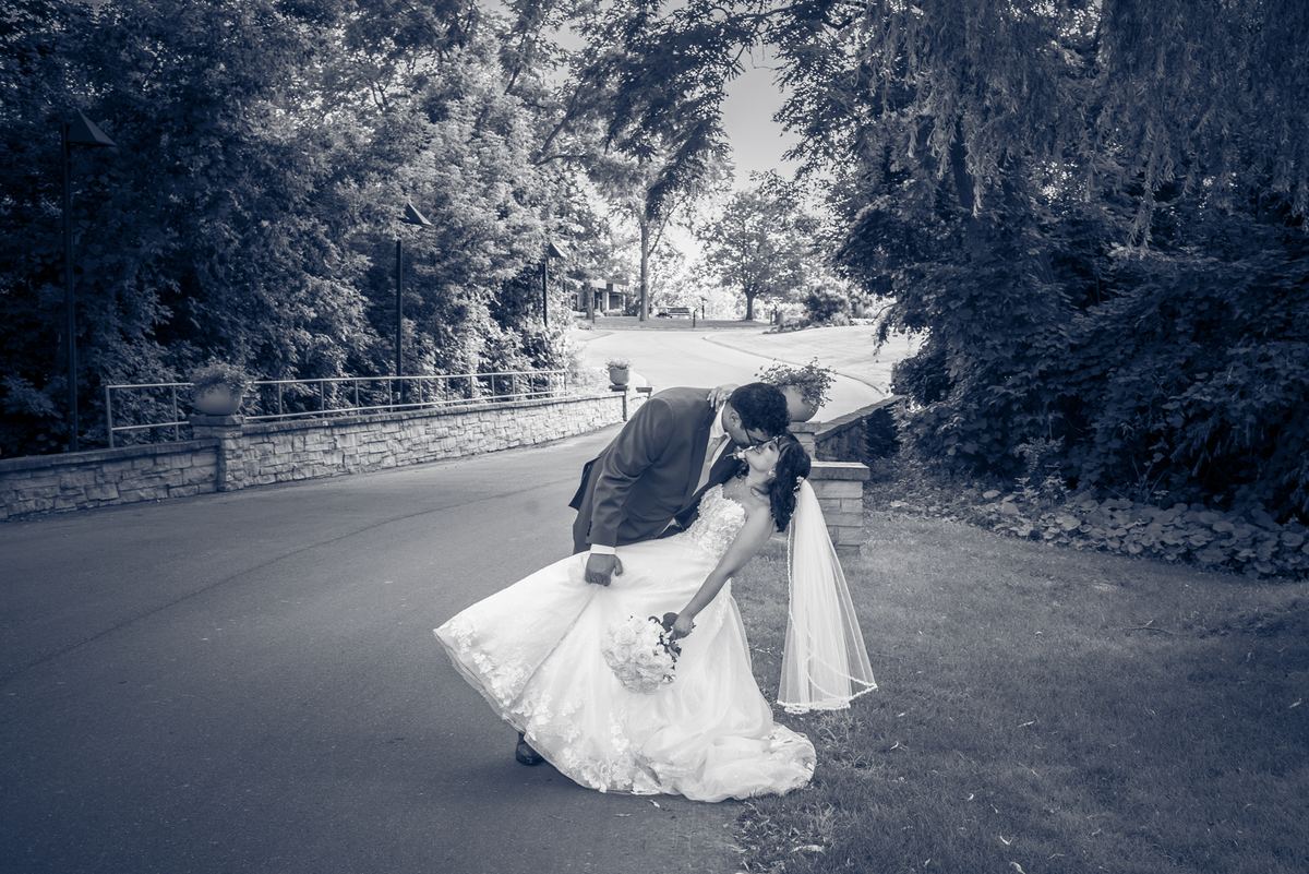 A bride and groom share a playful dip in an outdoor setting with lush greenery and a stone wall, bathed in soft daylight.