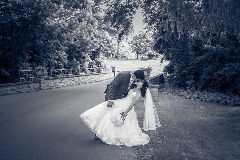 A bride and groom share a playful dip in an outdoor setting with lush greenery and a stone wall, bathed in soft daylight.