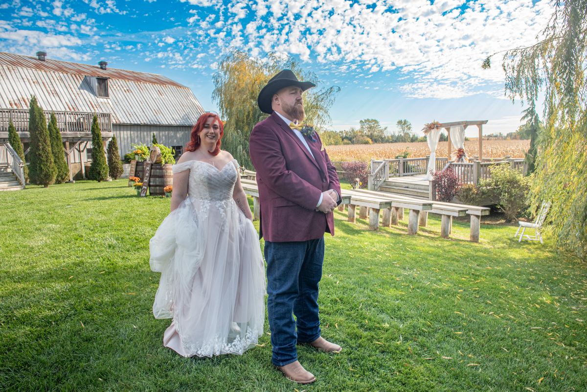 A red-haired bride and groom smile at each other on a grassy lawn during an outdoor wedding ceremony setup.