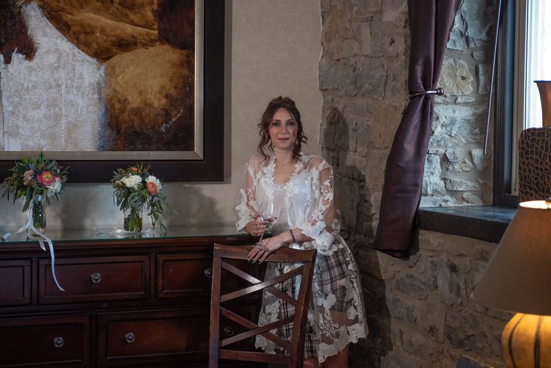 A woman in lace and plaid stands by a wooden chair in a rustic room with stone walls, holding a wine glass.