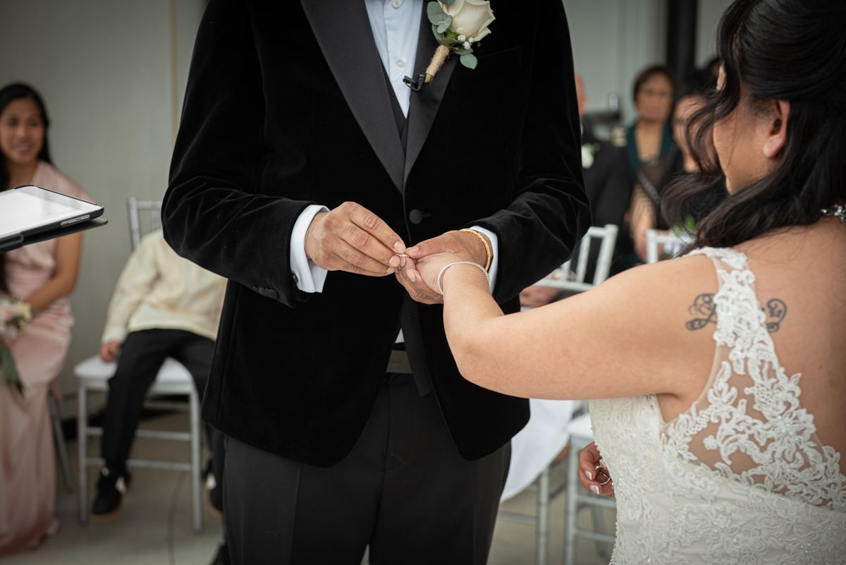 Groom in velvet tuxedo places ring on bride's finger as she extends her hand, both surrounded by blurred guests.