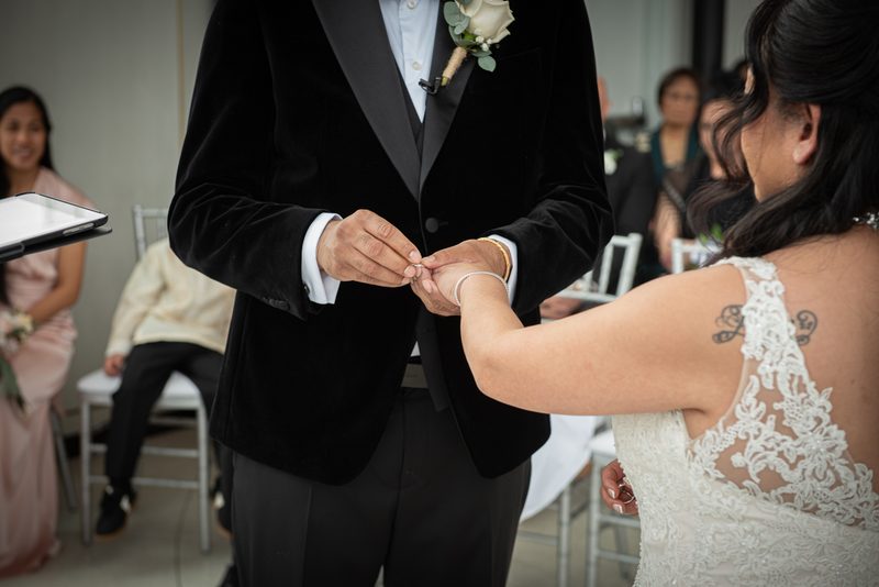 Groom in velvet tuxedo places ring on bride's finger as she extends her hand, both surrounded by blurred guests.