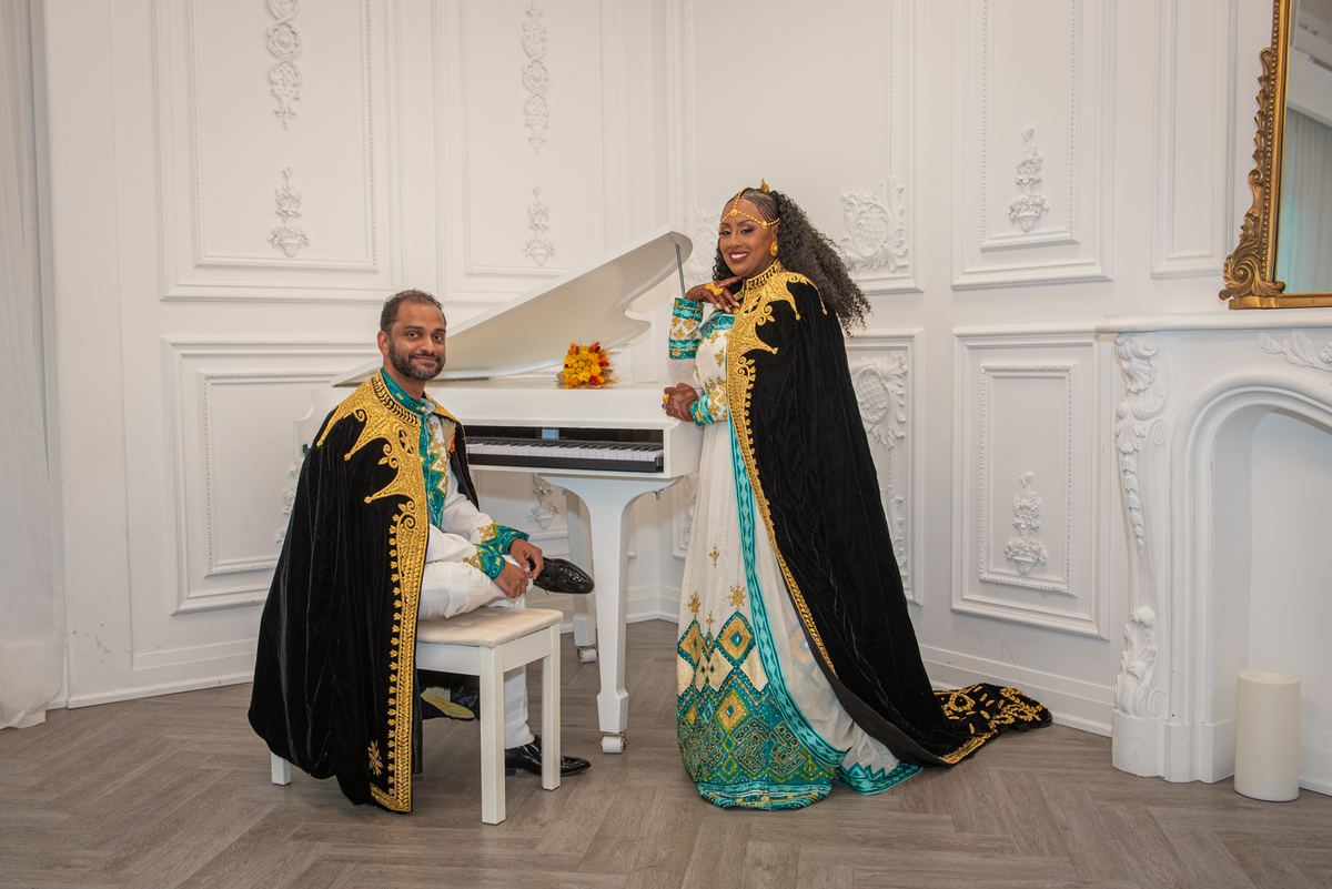 A man and woman in traditional ceremonial attire stand beside a grand piano in a white-walled room, smiling.
