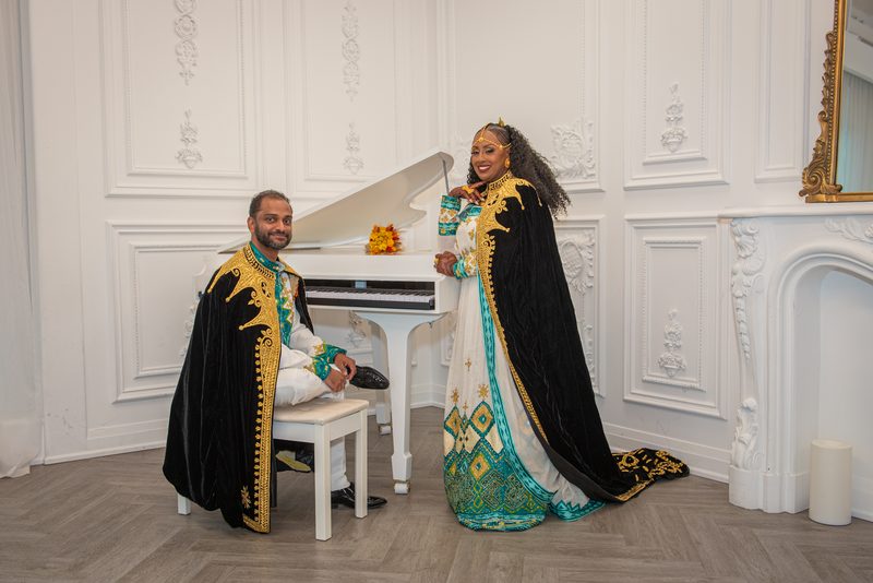 A man and woman in traditional ceremonial attire stand beside a grand piano in a white-walled room, smiling.