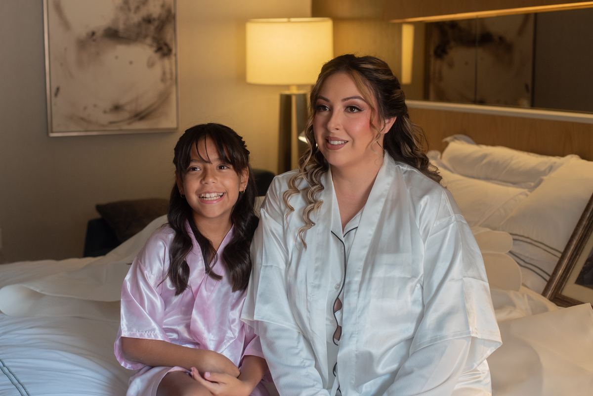 An adult woman and a young girl sit together on a hotel bed, both smiling warmly at the camera in matching satin robes.