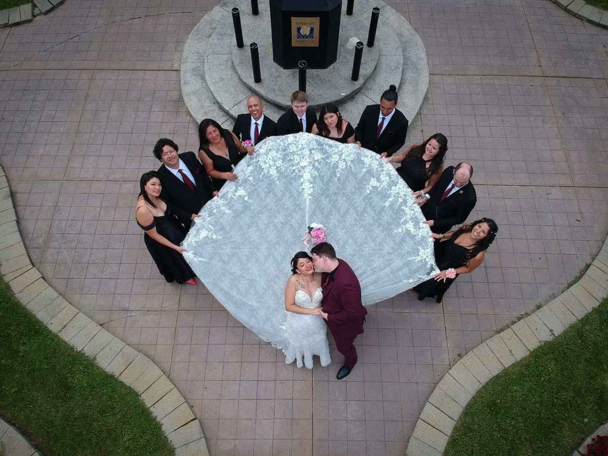Bride and groom embrace under wedding party's lifted veil, forming an umbrella canopy around them on a plaza.
