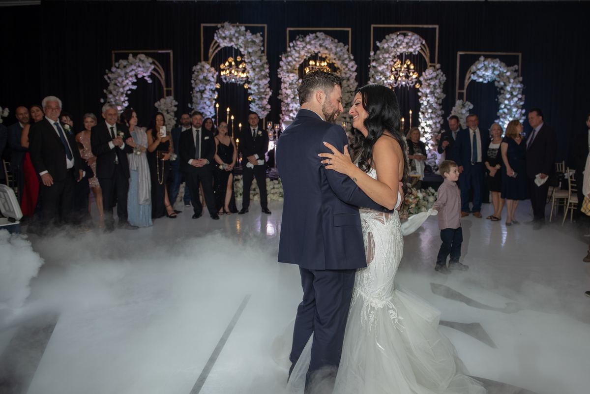 A newlywed couple dances intimately as guests smile and clap in the background, illuminated by soft lighting.