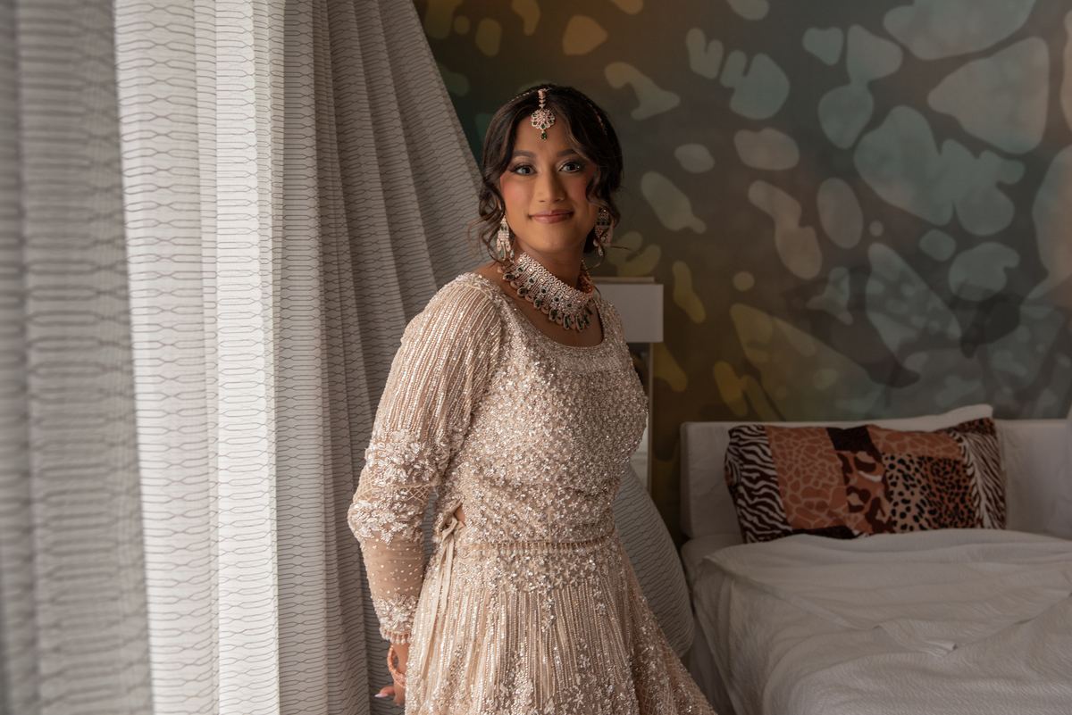 A bride smiles beside sheer curtains in soft natural light, with a patterned bed and textured wall behind her.