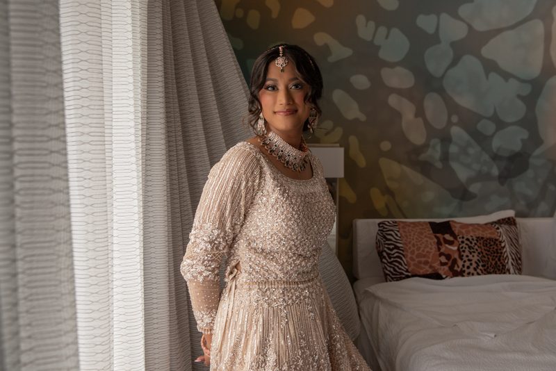 A bride smiles beside sheer curtains in soft natural light, with a patterned bed and textured wall behind her.