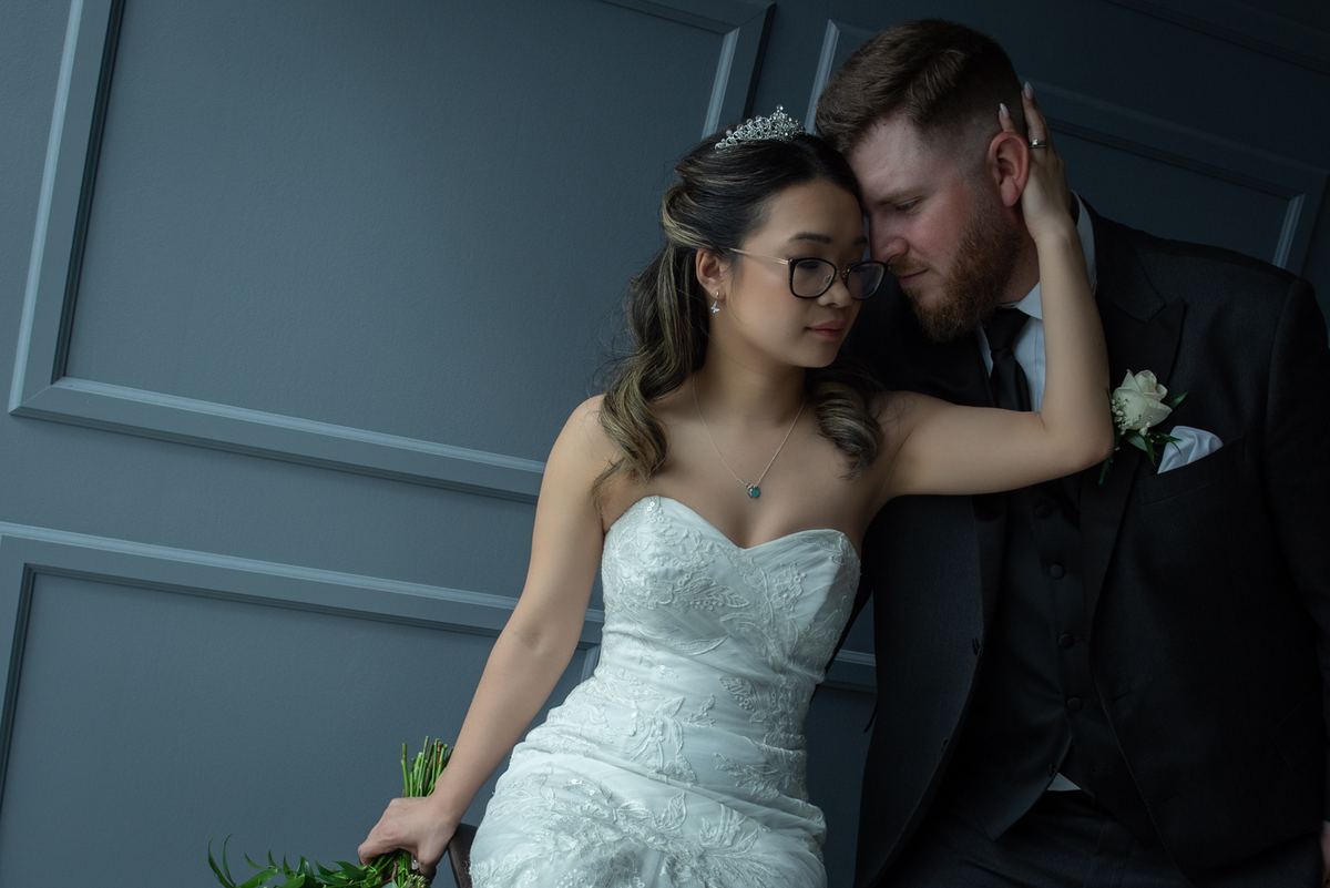 A bride in a white gown and tiara leans her head against her groom's, who wears a black suit, illuminated by soft light.