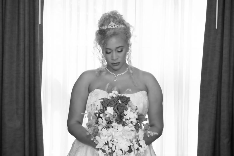 Bride in white dress and tiara holds bouquet, gazing thoughtfully downward with soft backlighting creating an ethereal glow.