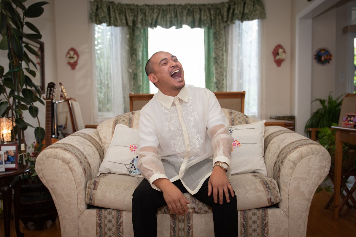 A man laughing on a patterned sofa in a cozy living room with green curtains and musical instruments.