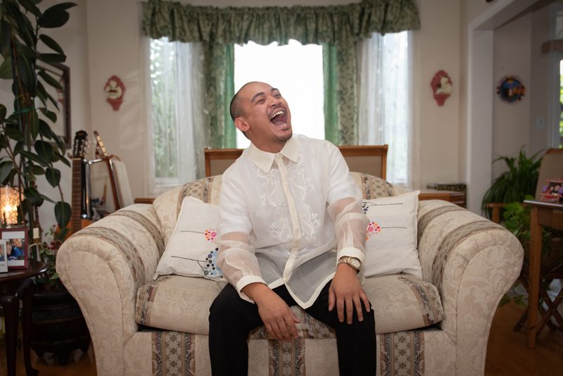 A man laughing on a patterned sofa in a cozy living room with green curtains and musical instruments.
