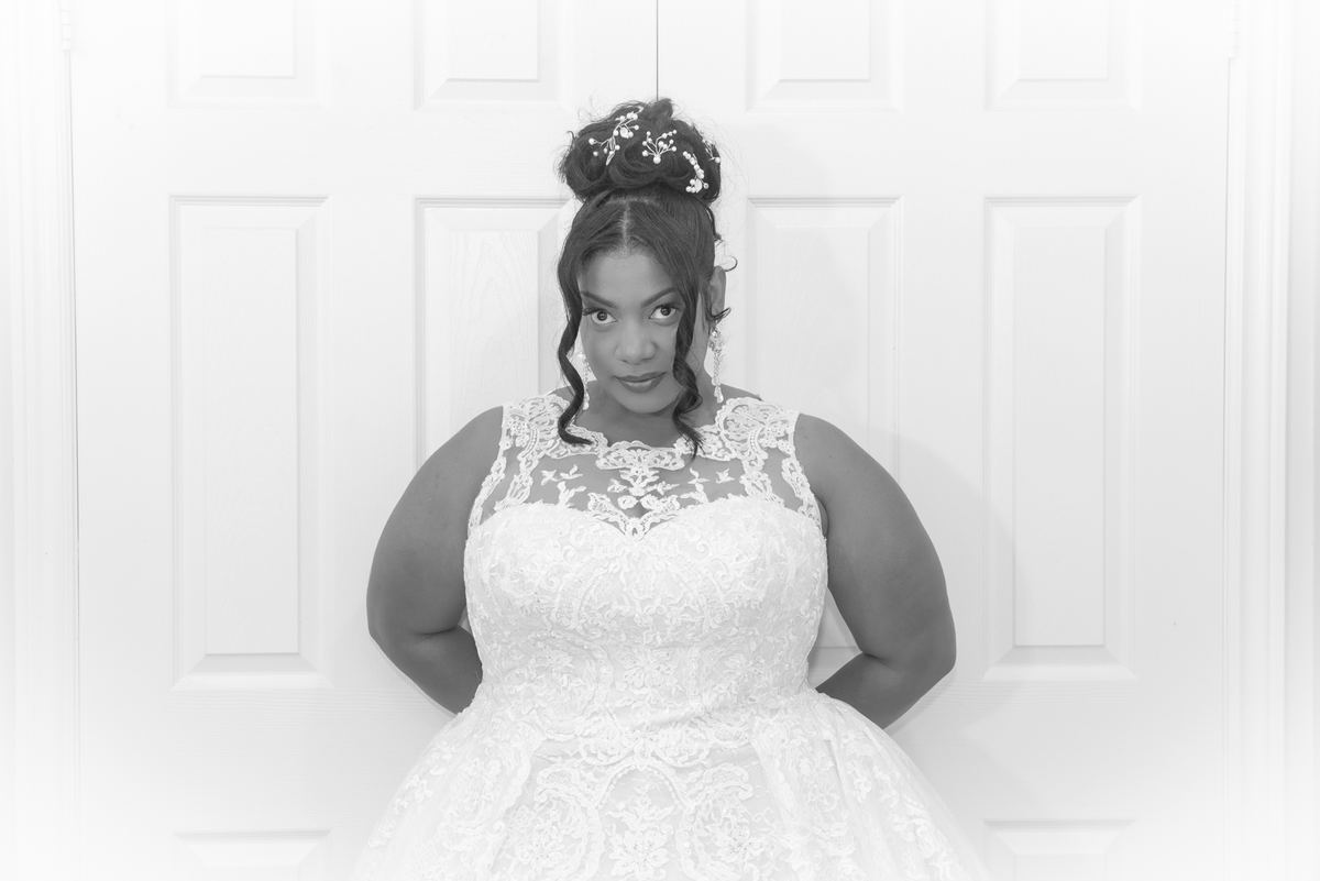 A bride in a lace gown stands confidently by a white door, hands on hips, hair adorned with delicate accessories.