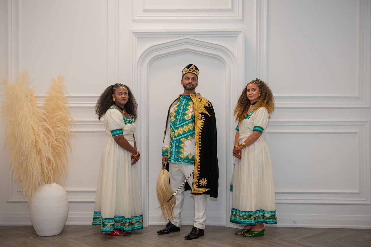 Three formally dressed figures pose against a white wall with an arched doorway and pampas grass arrangement, studio setting.