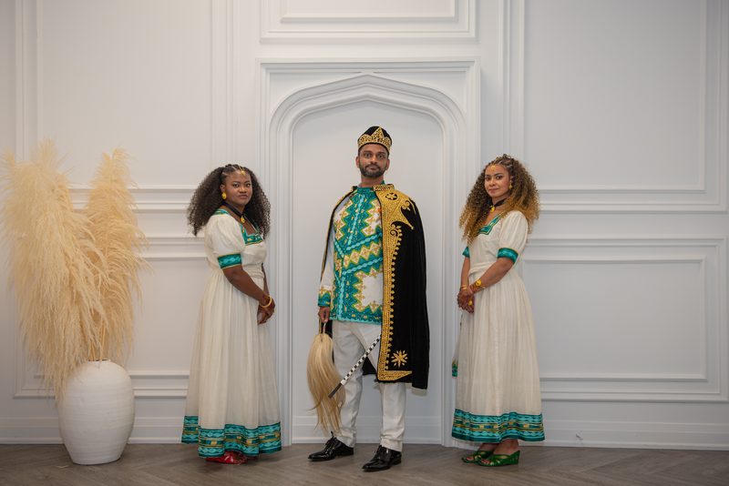 Three formally dressed figures pose against a white wall with an arched doorway and pampas grass arrangement, studio setting.